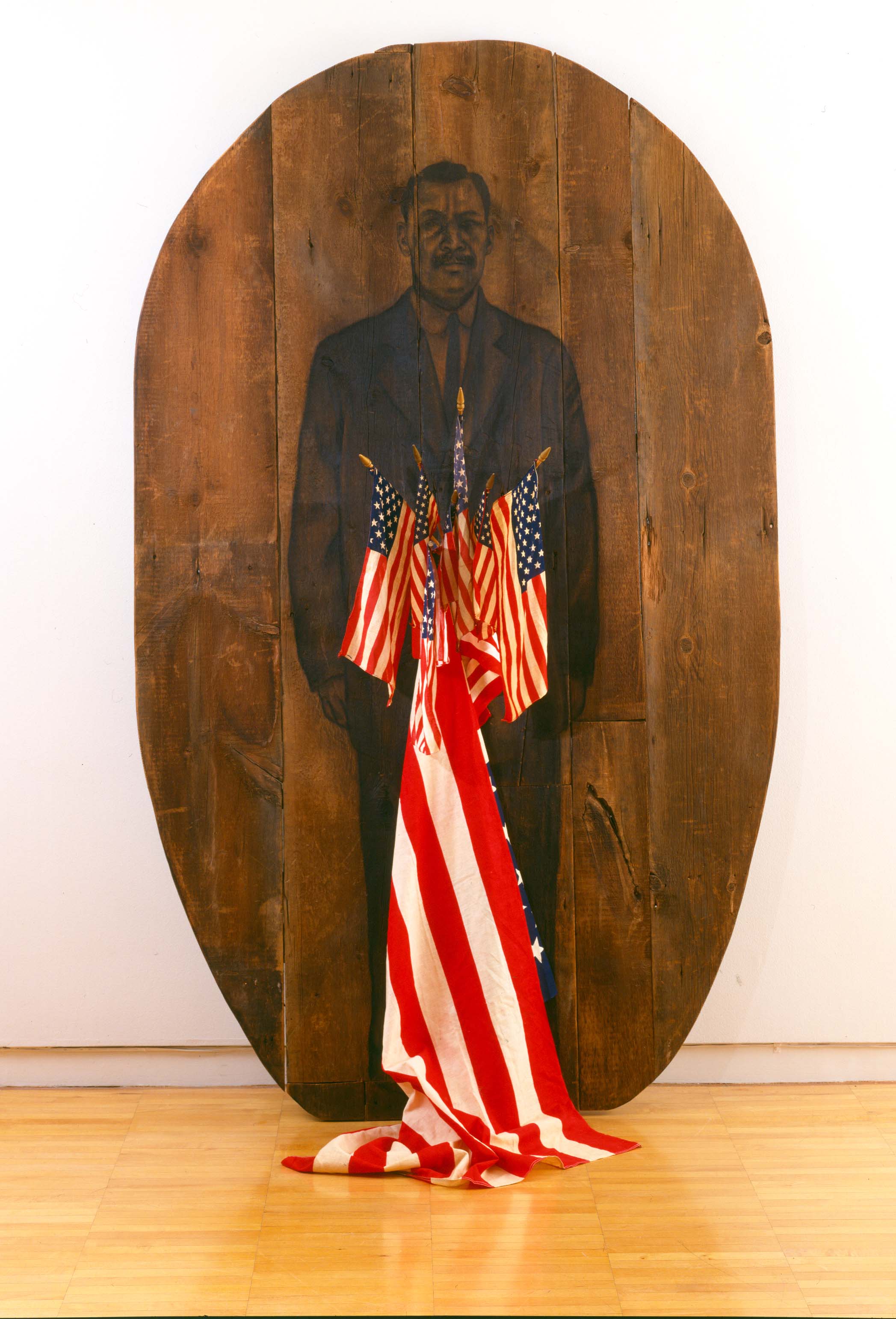 Large oval painting on wood planks of a Black man in a suit. American flags protrude from the front of the painting.