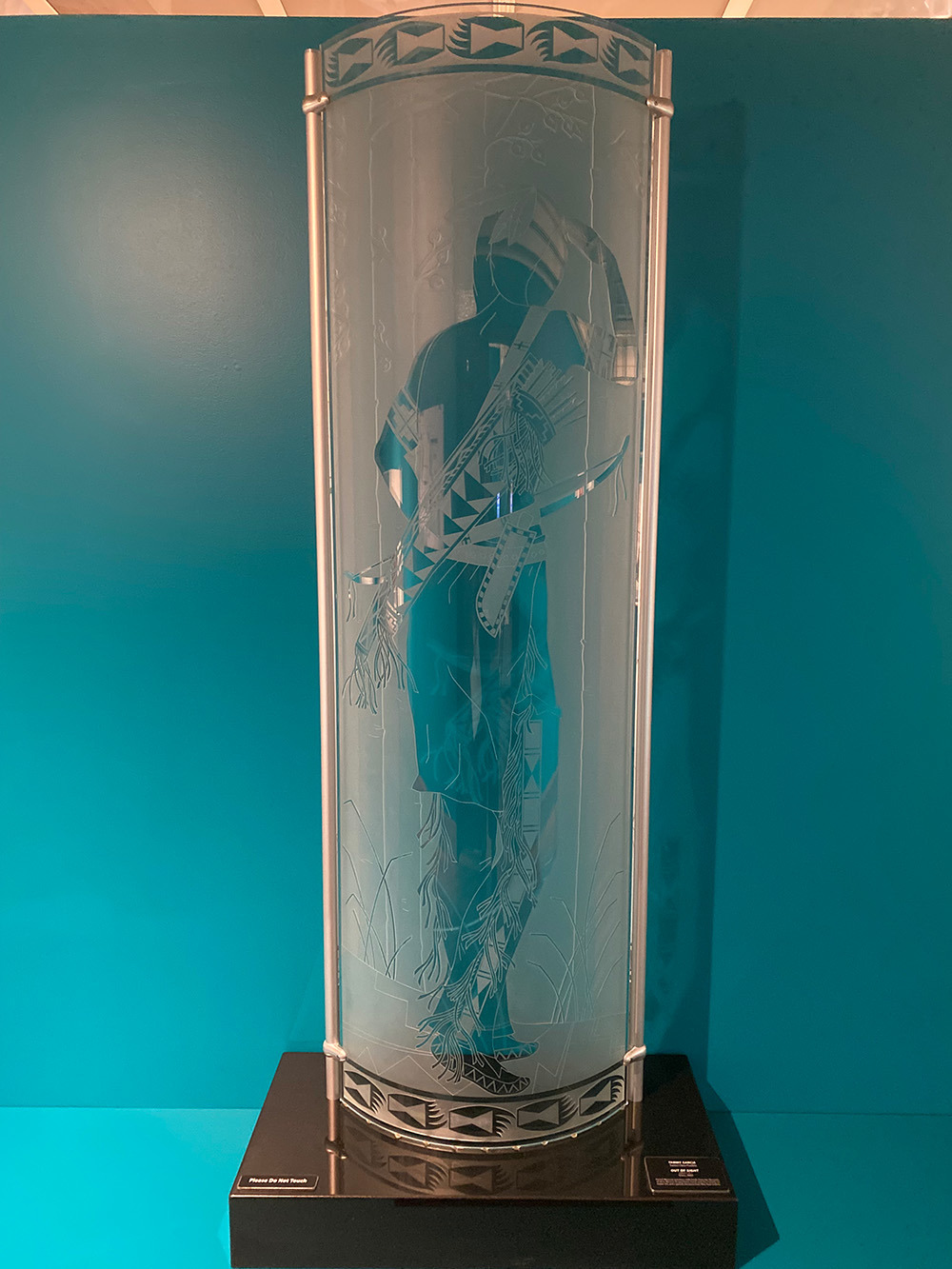 Out of Sight is a slumped and etched glass sculpture. A tall, rectangular convex glass panel is anchored to a black base by two vertical steel rods that extend from the top to the bottom sides of the panel.