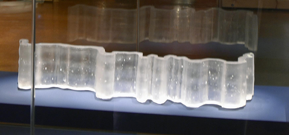 Pa’huk Site is a three-dimensional sculpture crafted from colorless cast glass. This artwork appears as a broad and crooked rectangular band, with bends and dips throughout, reminiscent of a flowing ribbon or the path of a curving river.