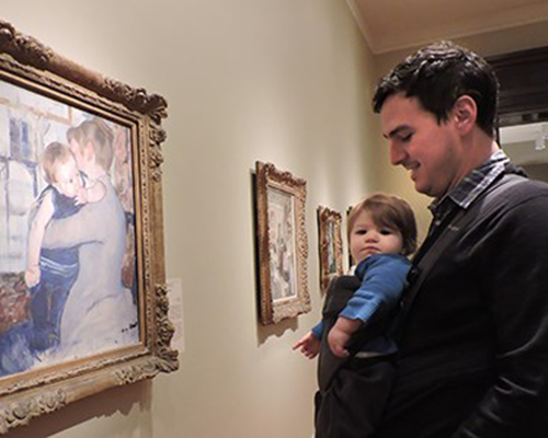 A young child in a sling looks at art with their parent