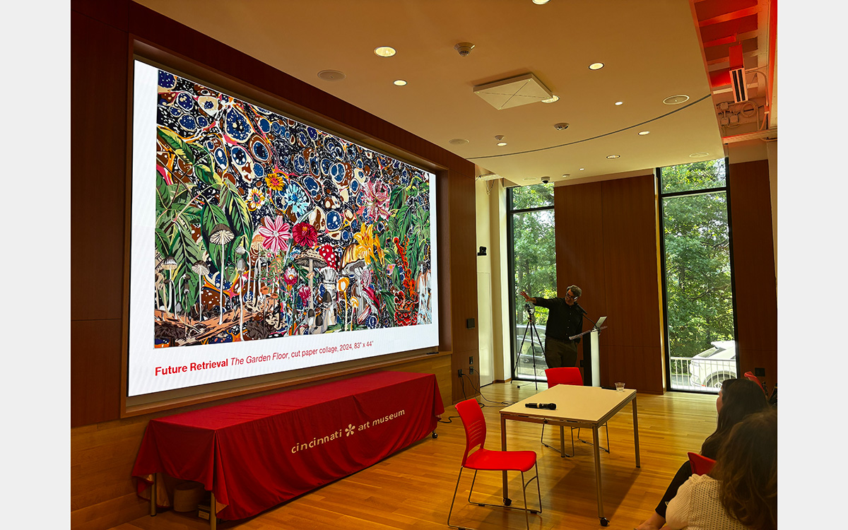 A man stands at a podium pointing to a large-scale presentation screen behind him.