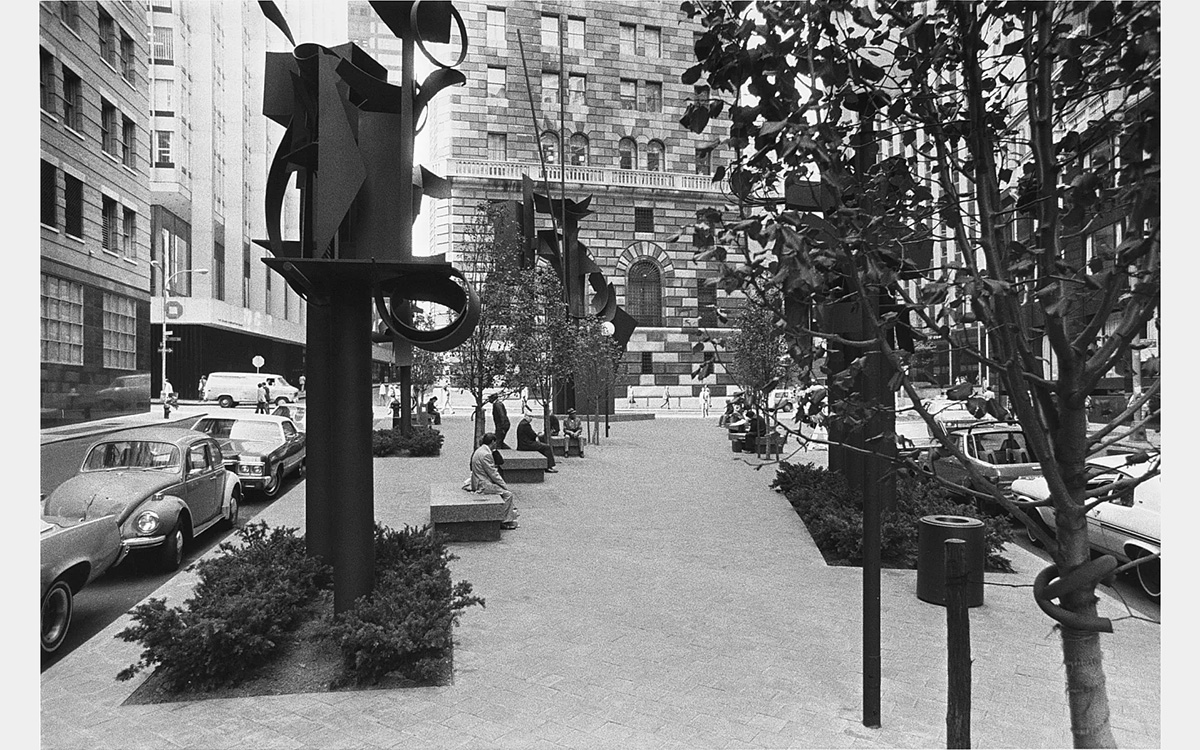 Urban park with sculptures and trees surrounded by parked cars and buildings