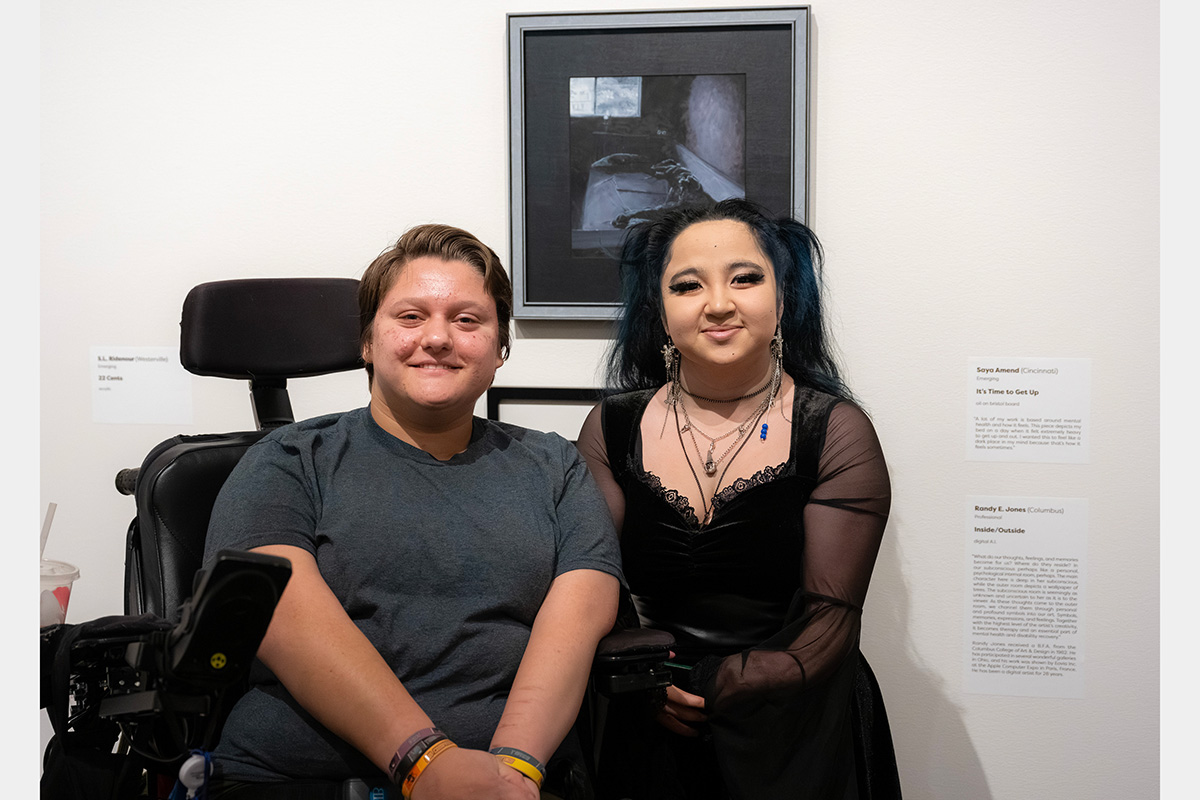 Two people smile in front of an artwork. One is sitting in a wheelchair