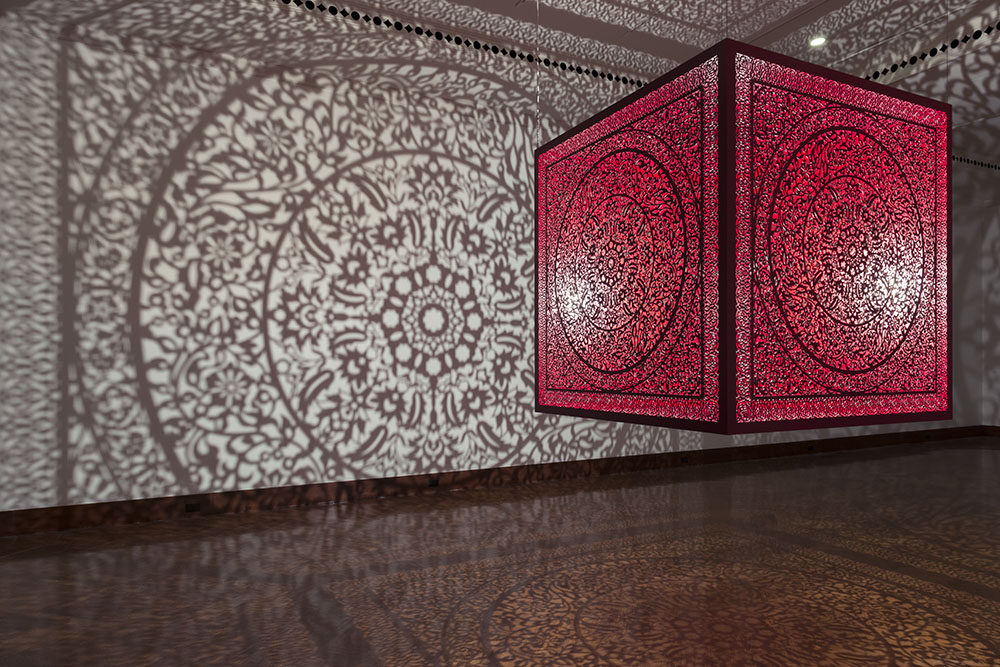Anila Quayyum Agha's All the Flowers Are for Me (Red), a large, red cube with a densely packed floral pattern carved into each side. A light source within the cube projects the pattern onto the walls, floor, and ceiling