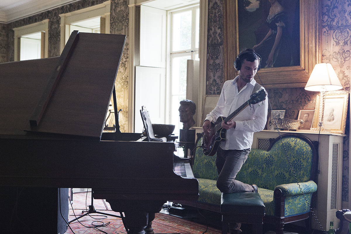 Photo graph of a man in a large living room, standing in front of a grand piano, playing a bass guitar