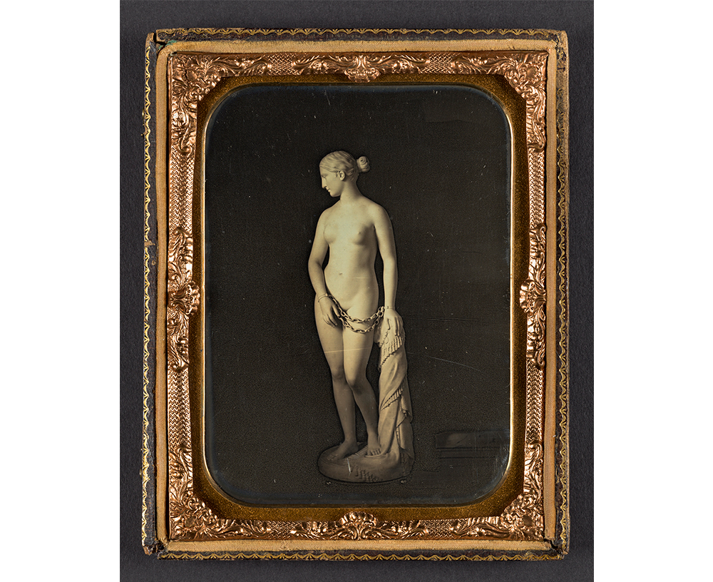 Abel Fletcher's The Greek Slave of Hiram Powers, an old sepia toned photograph of the front view of a marble statue of a nude woman wearing hand cuffs, her turned towards her right, and resting her left hand on a short post covered in cloth