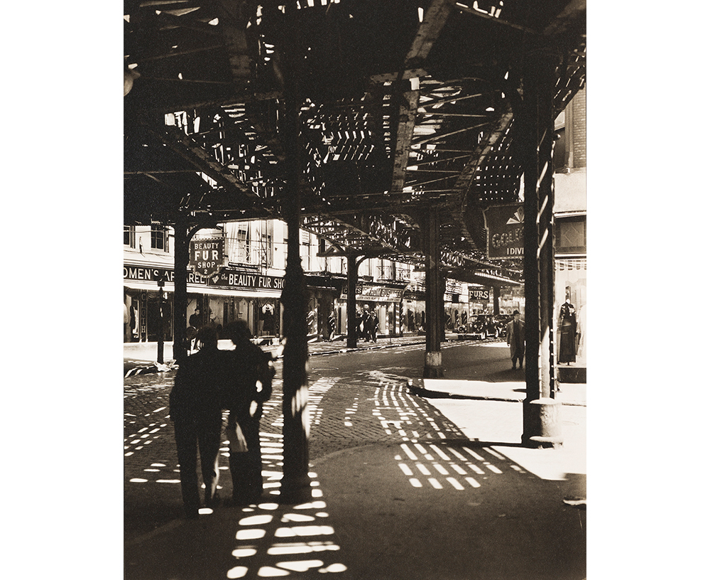 Berenice Abbott's El, Second and Third Avenue Lines, Bowery and Division Street, Manhattan, a sepia toned photograph of a New York City street beneath an iron train line. Pedestrians walk about the sidewalks past rows of store fronts and a parked car in the distance