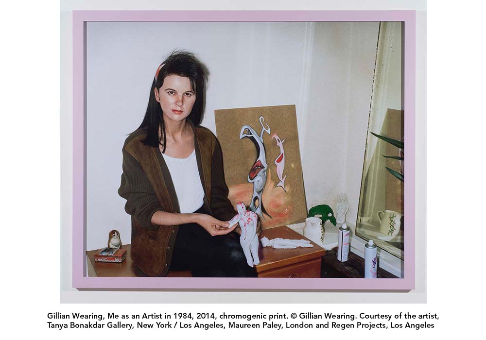 Gillian Wearing's Me as an Artist, A photograph of Gillian Wearing leaning on a desk in front of an abstract painting holding human figurine 