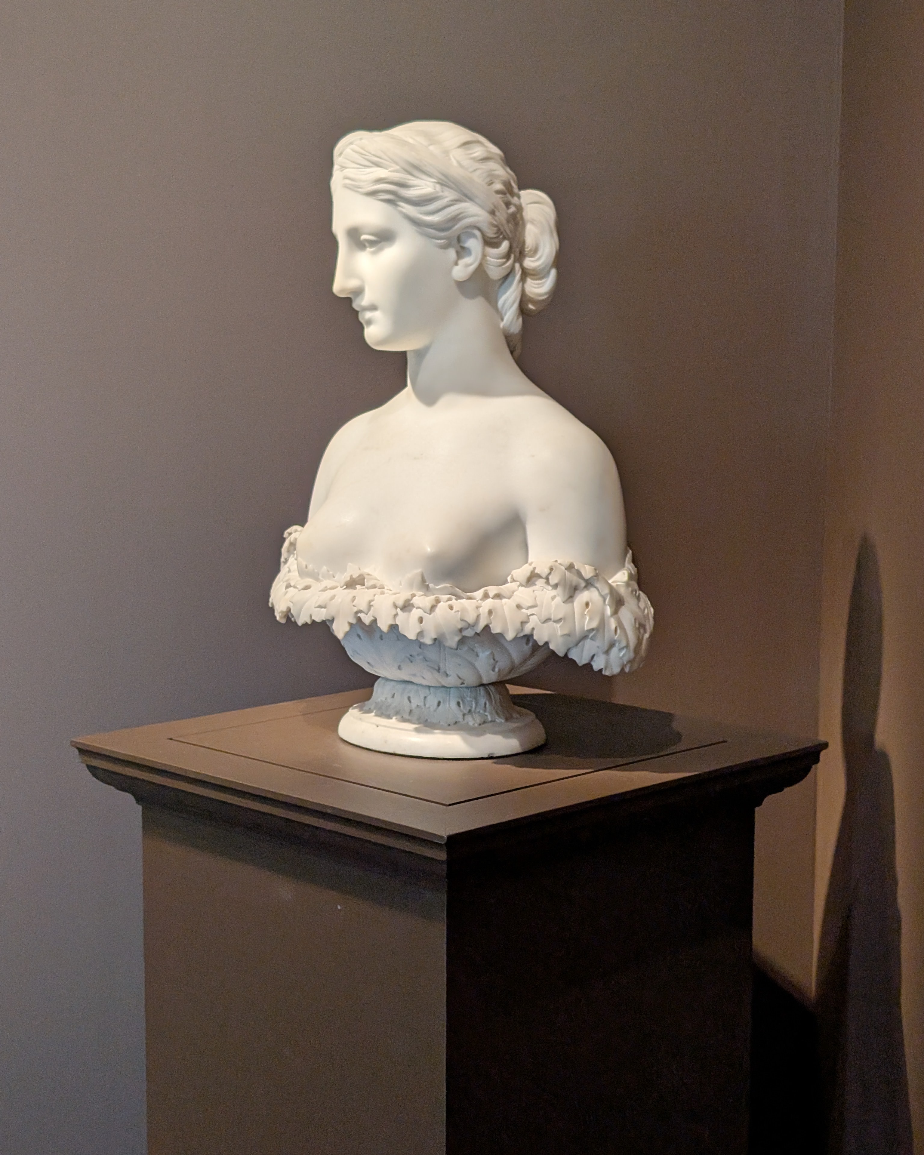 A white marble bust of a woman sits on a brown pedestal against a light brown background.