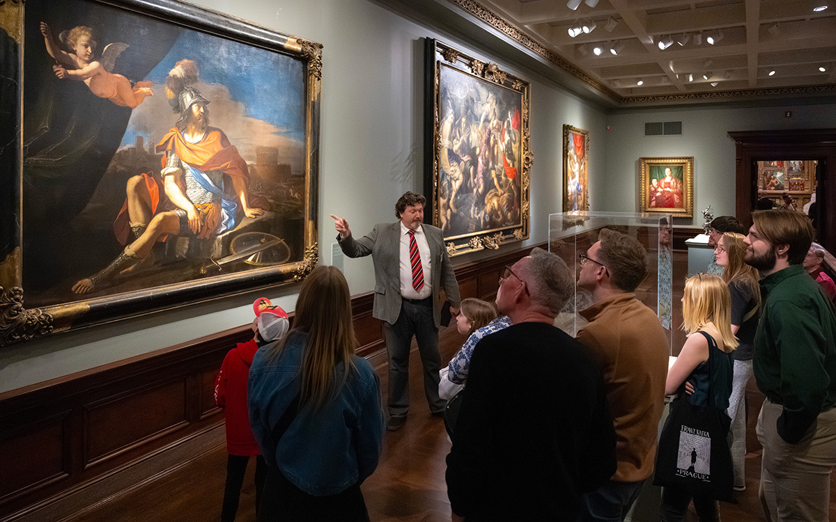 A docent points at a massive painting in front of a crowd of people