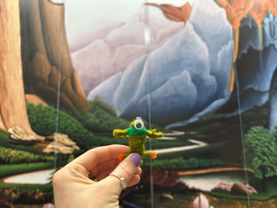 A human hand holds an animal-like figure made of yard against the backdrop of a landscape painting.