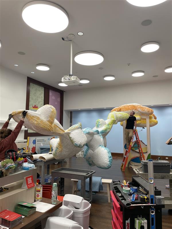 Installers -- one standing on a ladder -- hold a long line of cloud-like forms in preparation to mount the forms to the ceiling.