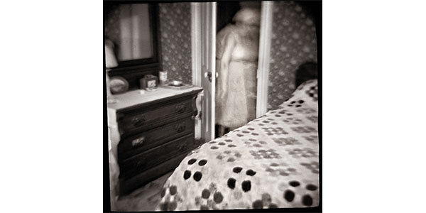 Nancy Rexroth's Clara in the Closet, black and white photograph of a bedroom, the blurry figure of a woman is walking through a door in between a dresser and a bed
