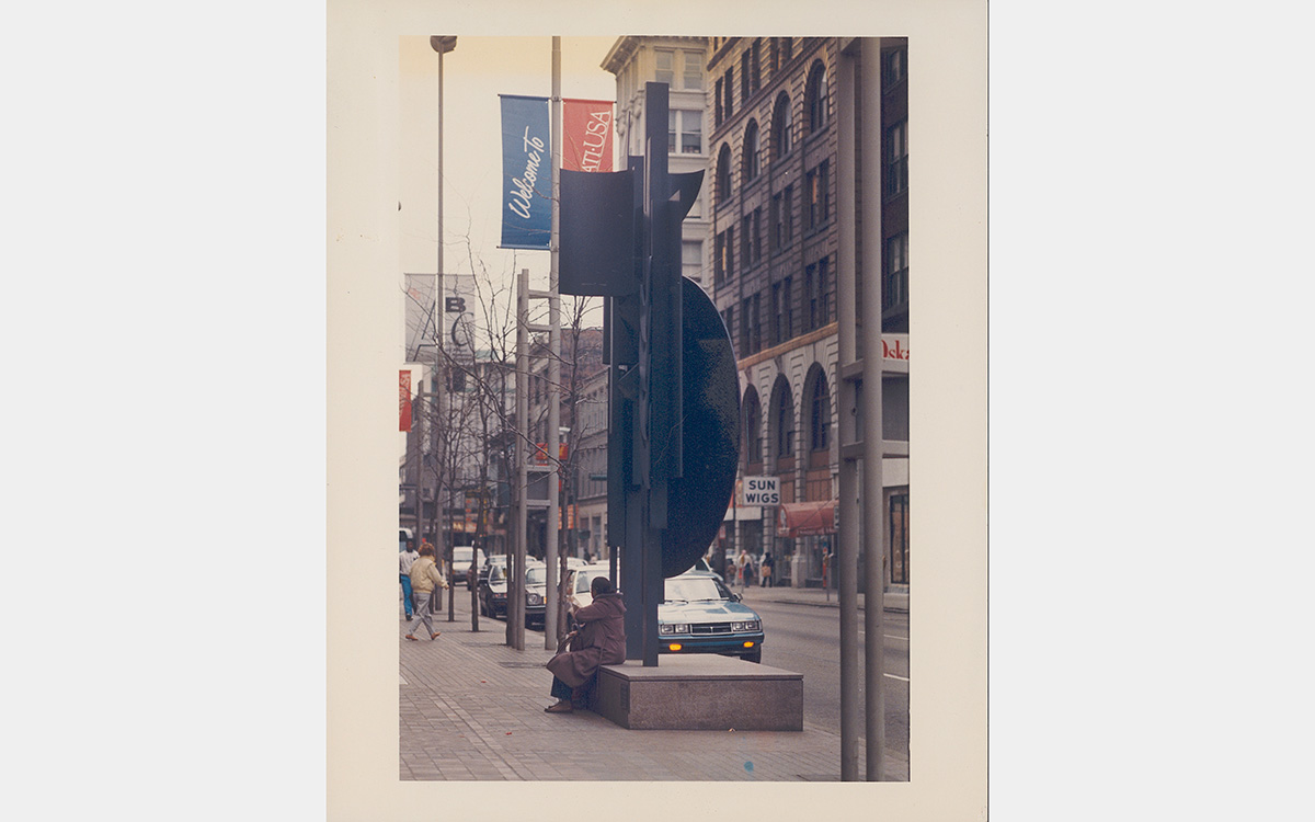 Faded color photo of person sitting on the base of a black metal sculpture on a streetscape with buildings in the background