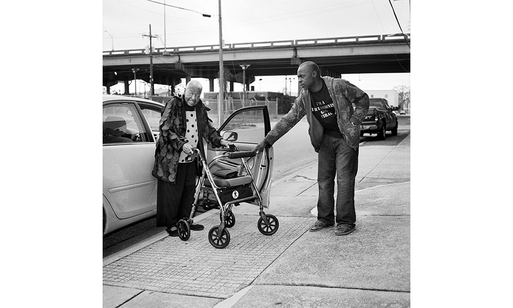 A black and white photograph of an African American man helping an elderly woman out of her car and onto the sidewalk. The woman holds tightly to a walker and smiles looking toward the photographer