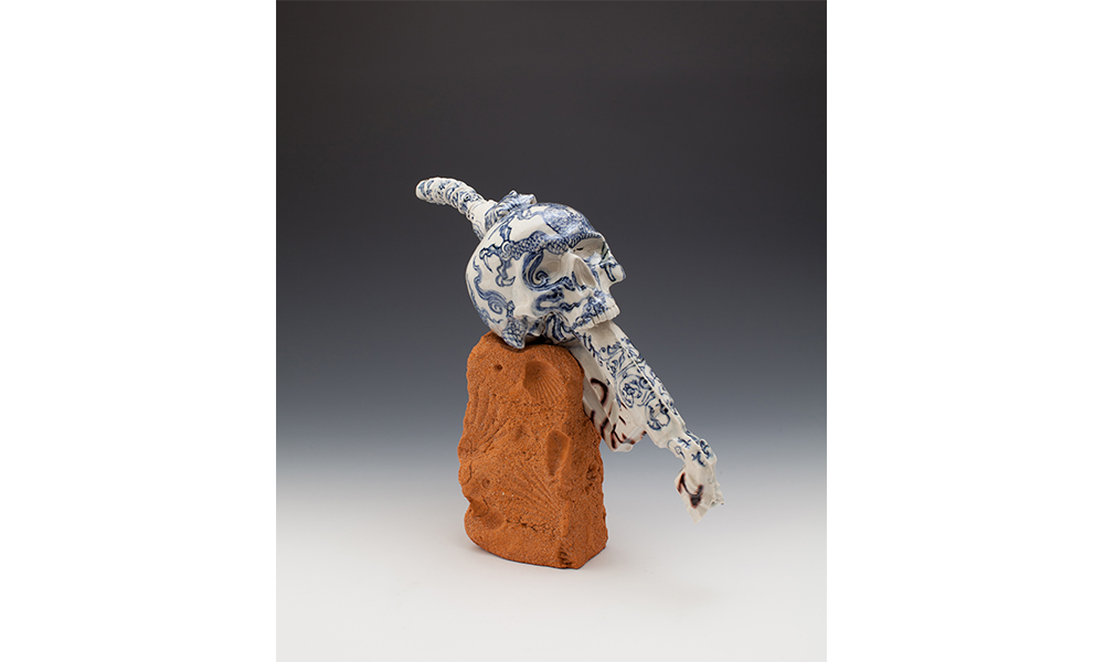 Michelle Erickson's Weapon of Choice, a porcelain statue of a skull embedded onto a fuel nozzle, decorated with a Chinese style dragon in blue glaze. The skull and nozzle rest on a brick impressed with fossilized shells