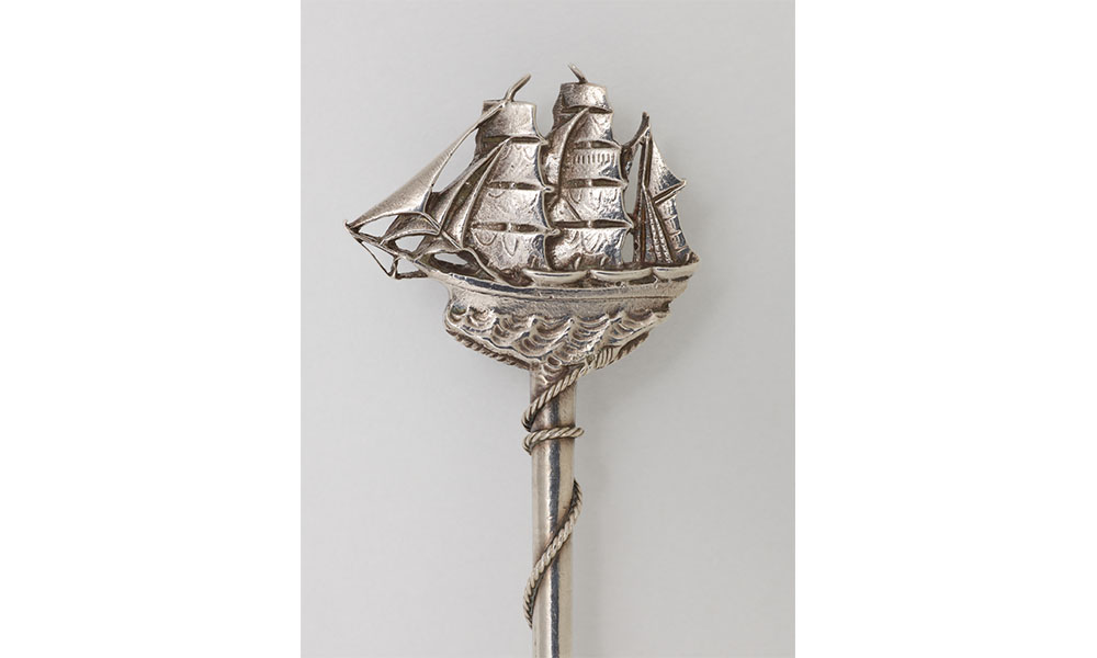 Handle of a silver teaspoon shaped like a large sailboat moving through rough waves