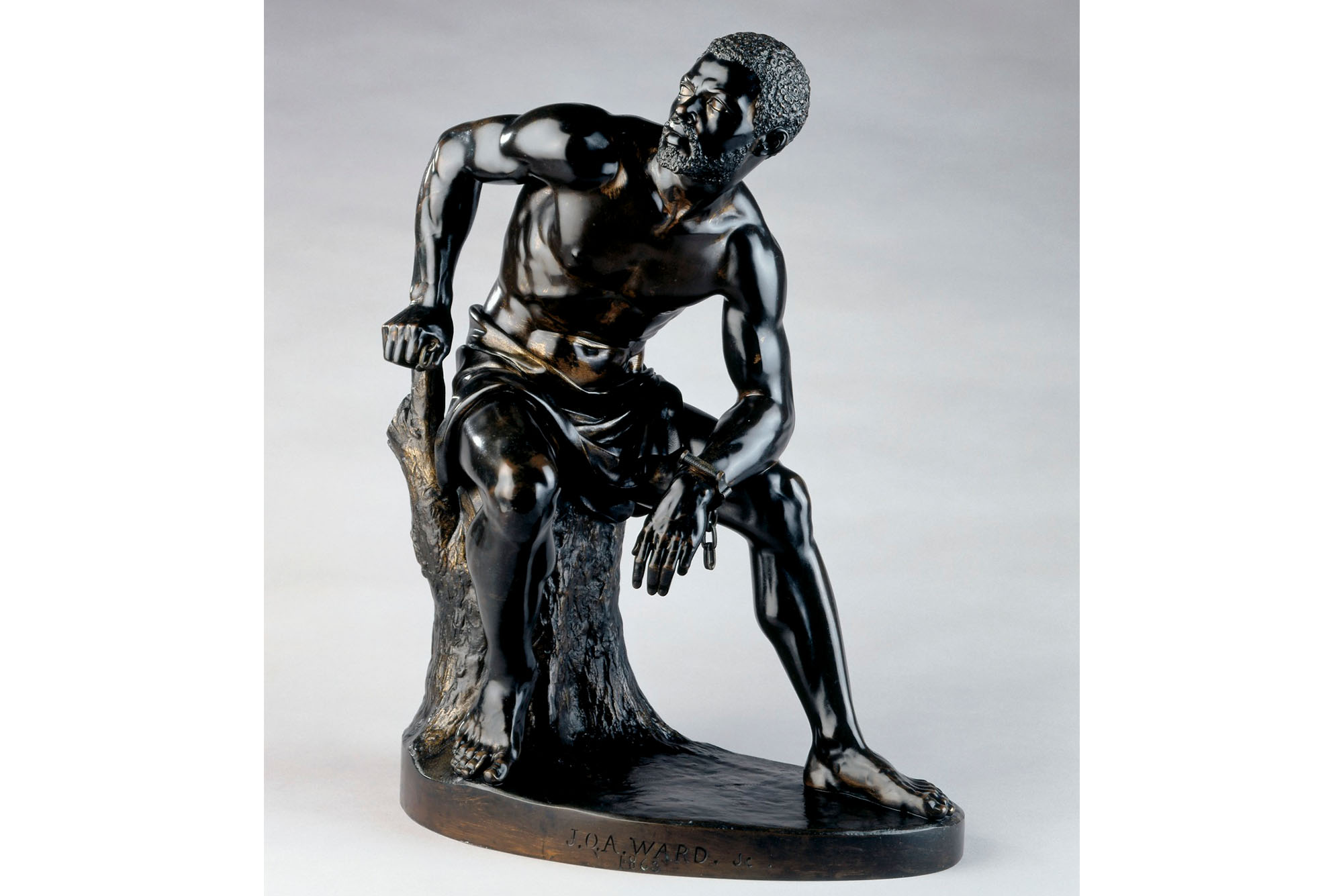 John Quincy Adams Ward's The Freedman, a bronze statue of an shirtless African American man sitting on a stump. He sits hunched over and turned slightly looking to his right with his right hand on the stump. On his left wrist is a single handcuff, the chain dangling down.