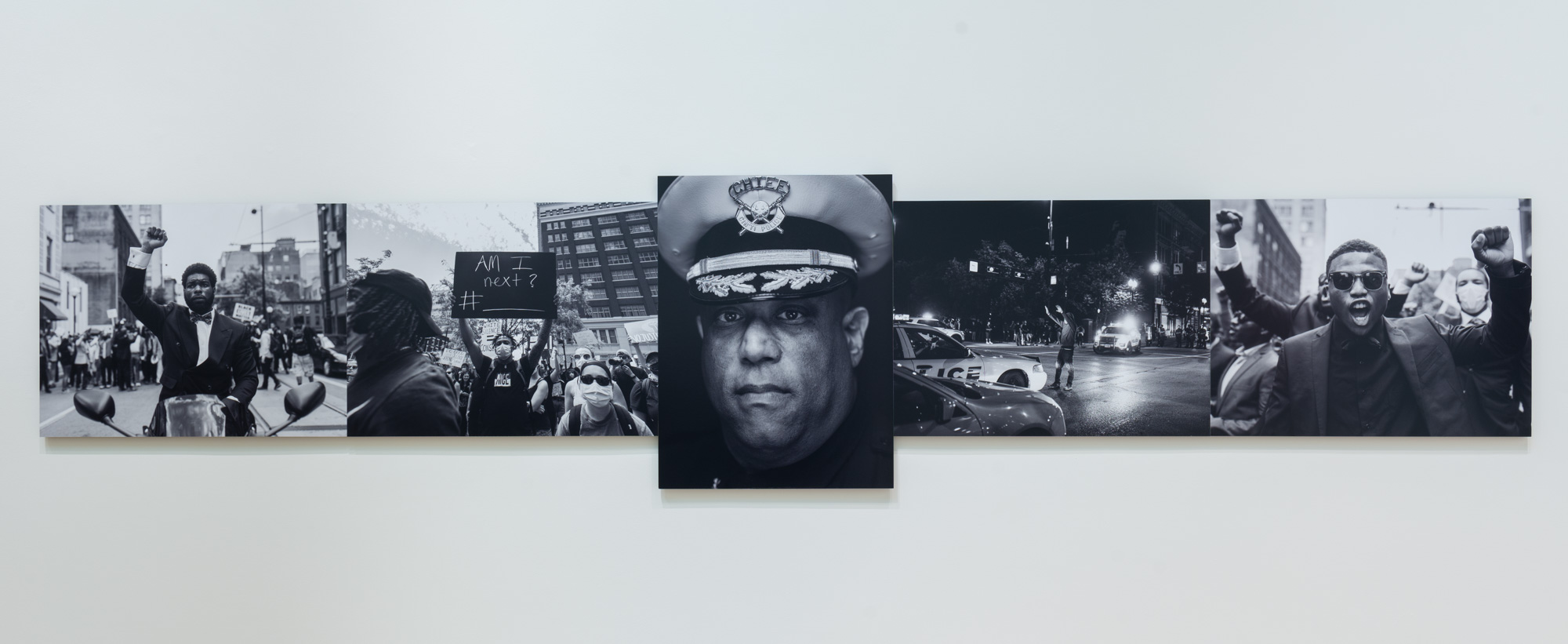 Kevin J. Watkins' Arrest the cops that killed (insert name), a series of black and white photographs of African Americans in protest. The center photograph is a close up portrait of an African American police chief in uniform