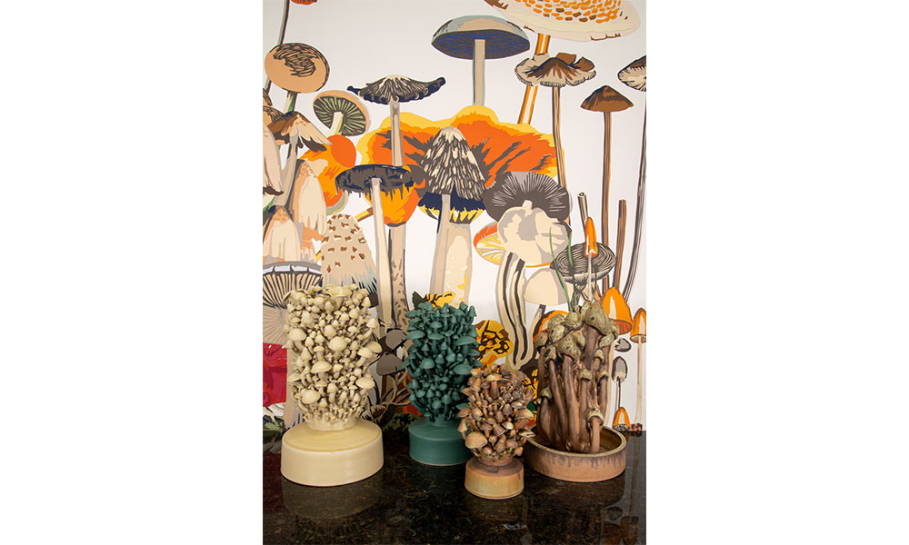Consolarium, white, green, and brown, porcelain statues of mushrooms places in front of hand cut paper mushrooms of various shapes, sizes, and brown-orange hues