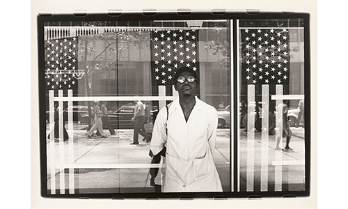 black and white photograph of an African American man standing in front of a window reflecting the New York City sidewalk, behind the window hang three American flag banners