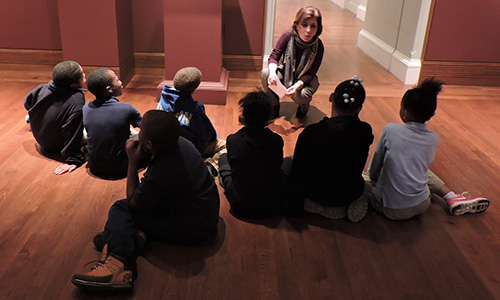A group of students sit in the gallery with their tour guide