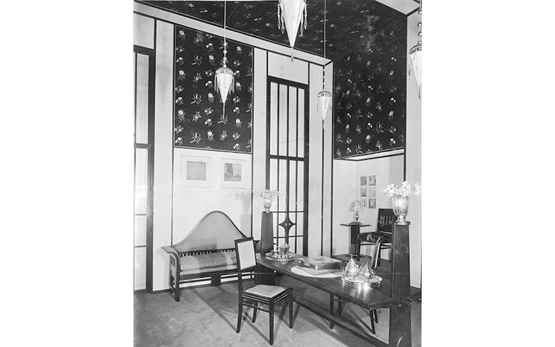 Black and white photo of an interior with dark furniture, dark floral wallpaper, a silver tea service and vase, and triangular hanging lamps. 