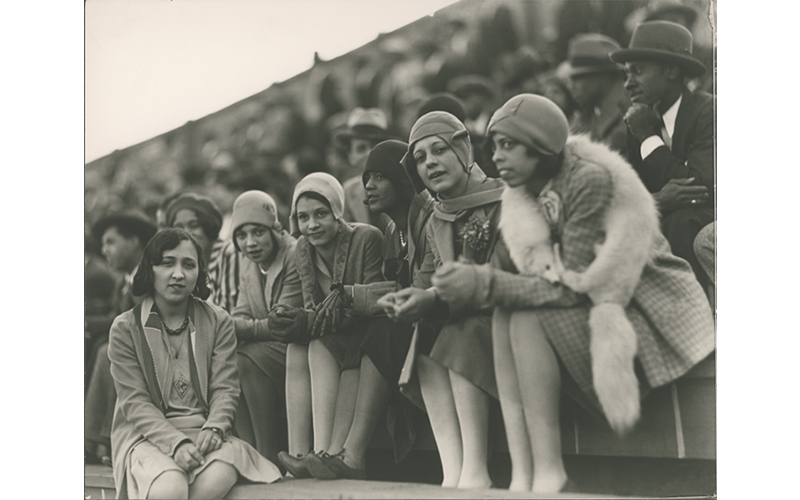 Six young women sit in a crowded stadium, with bobbed hair, cloches, raised hemlines, and scarves. Four of the six women look directly at the camera, the others look towards what is happening on the field.  