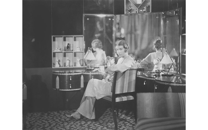 Young woman sitting at her mirrored dressing table facing the camera.