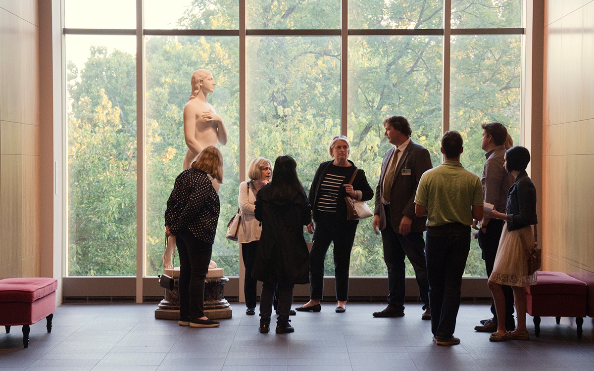 A group of people stand near a marble statue of the woman with a large window in the background overlooking green trees.