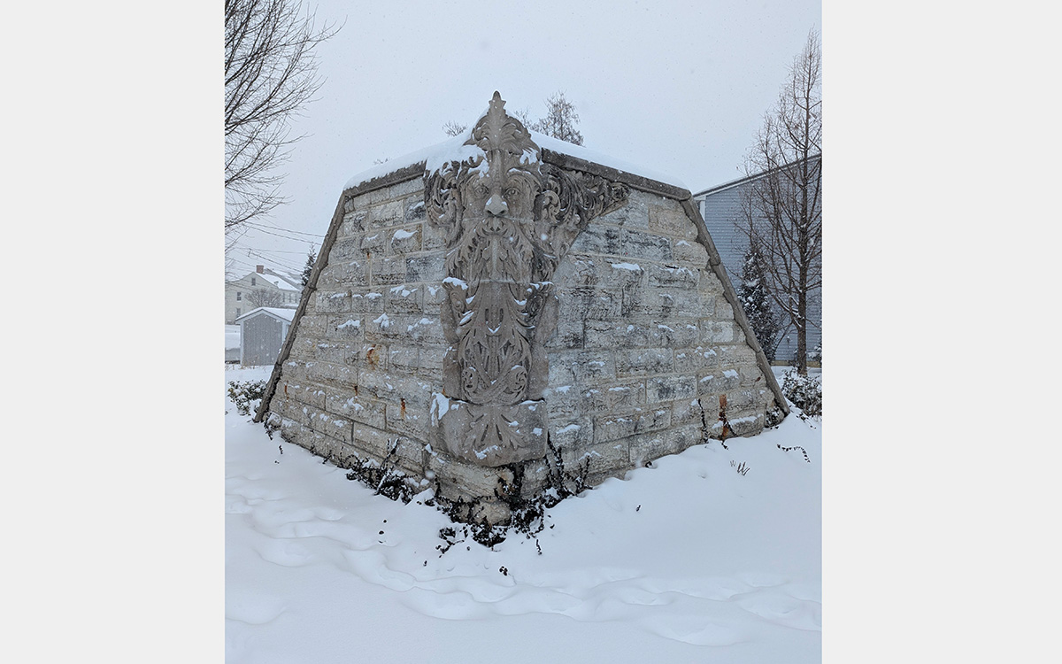 Two rough-cut stone walls intersect at right angles. At the corner is a stone carving of a bearded face with foliage projecting from around it.