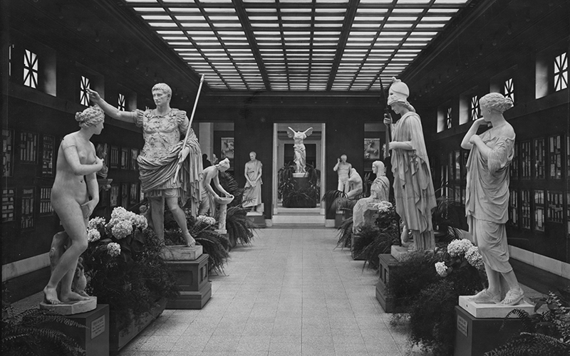 A historic black and white photo of a hallway full of sculptures of people