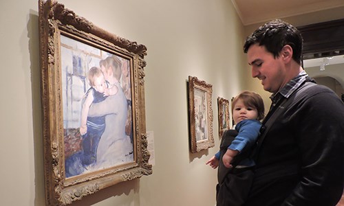 A baby in a sling and their parent enjoy an artwork