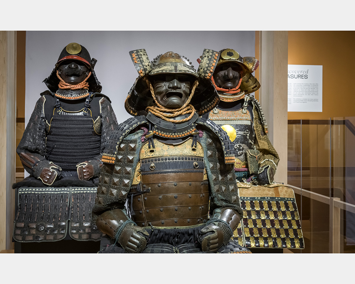 Three mannequins wearing suits of armor, helmets, and facemasks "sit" on platforms with a transparent barrier on the right.