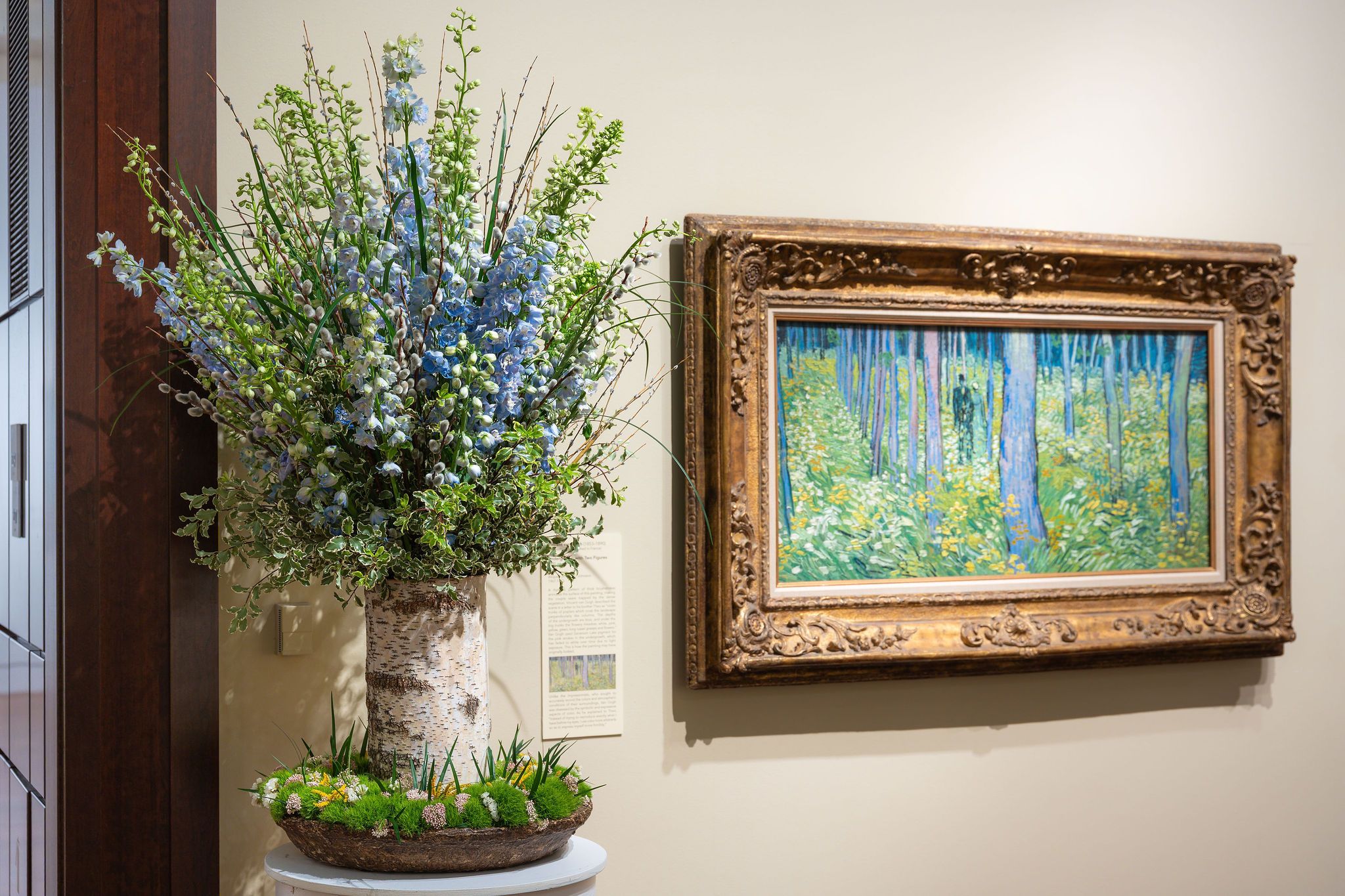 Large floral arrangement next to a colorful painting in a large gold frame.