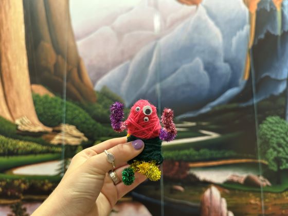 A human hand holds an animal-like figure made of yard against the backdrop of a landscape painting.