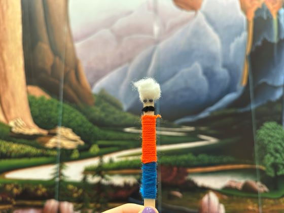 A human hand holds an animal-like figure made of yard against the backdrop of a landscape painting.