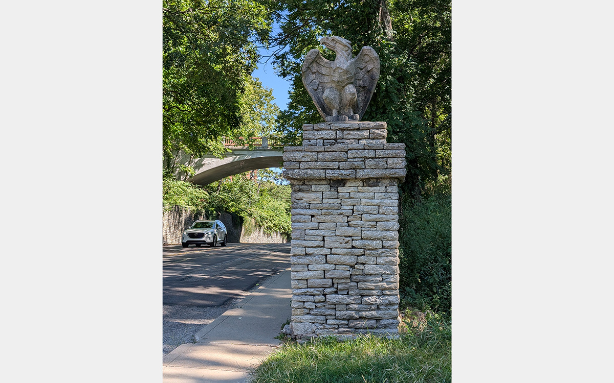 Carved stone eagle set atop a stone pedestal. In the background, a white car drives along a road under a bridge.