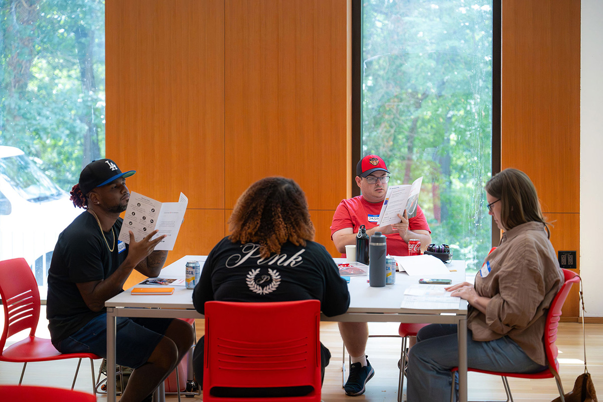 Four people sitting around an indoor table looking at brochures with views of trees through windows in background.