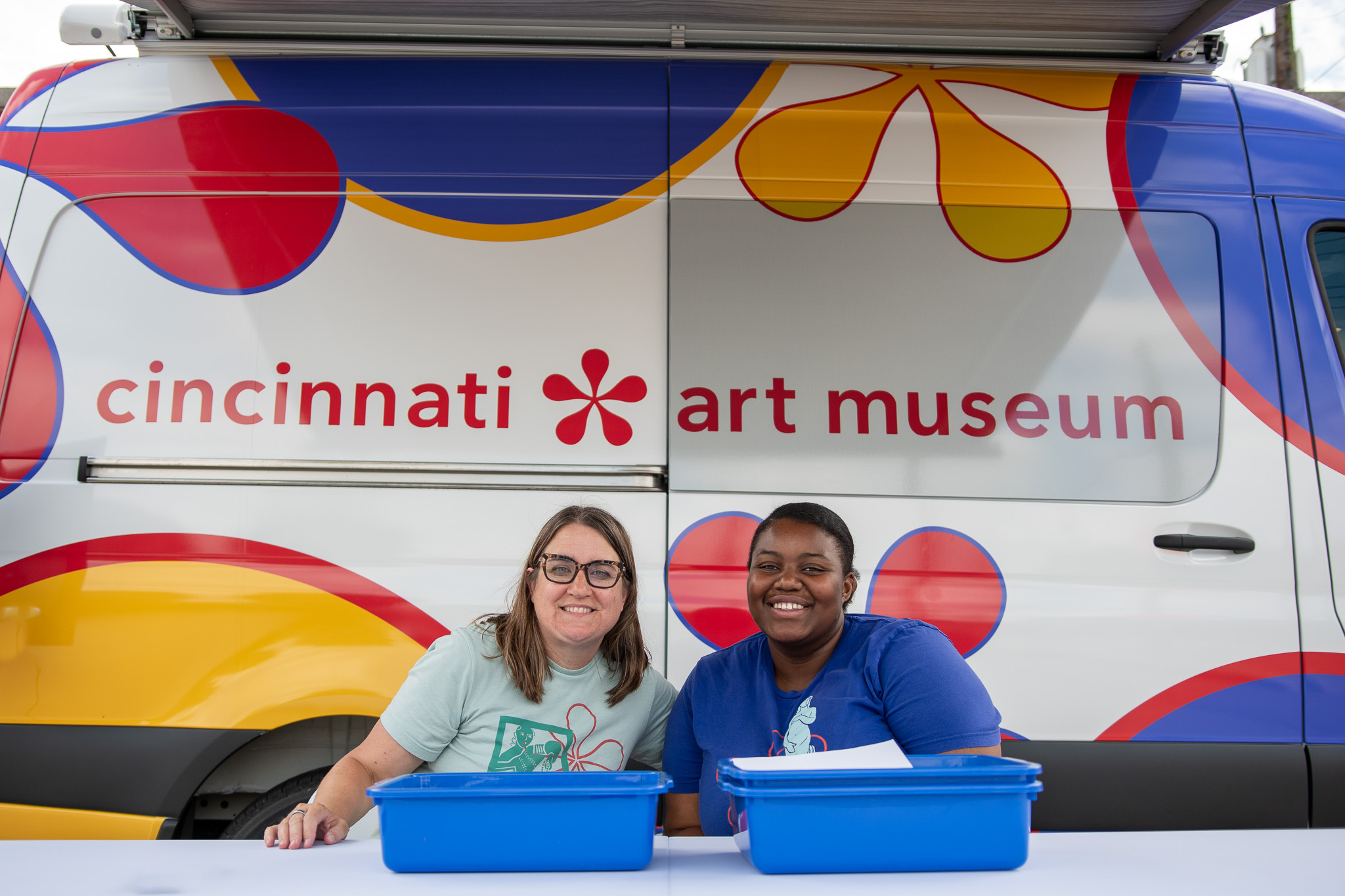 Two women sit at table outside of a brightly colored van with "cincinnati art museum" painted on the van's side.