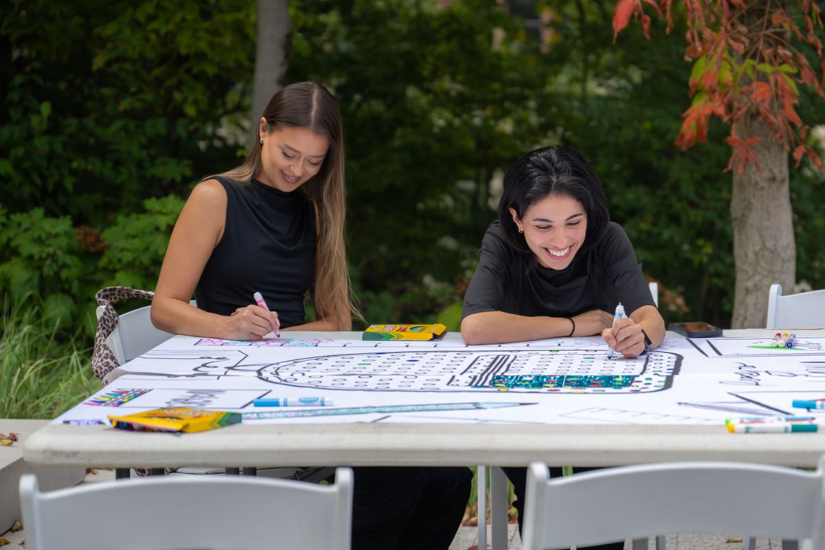 Two women coloring a black and white illustration on a table located in outdoor setting with green trees in the background.
