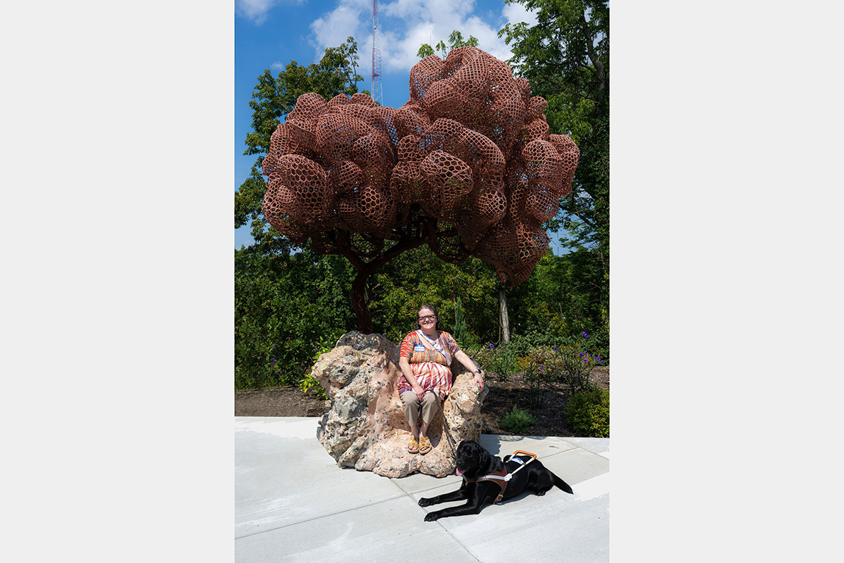 Woman seated under an outdoor sculpture of a tree with service dog laying on the ground.