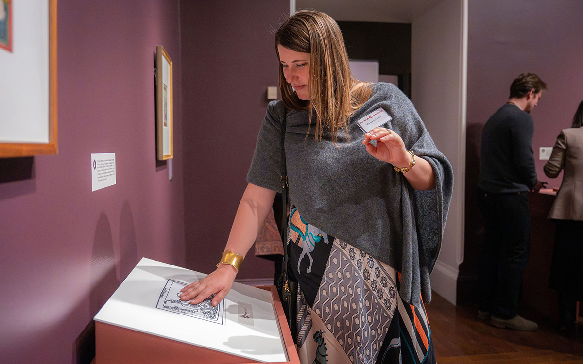 A woman places her hand on a touchable artwork installed on a tabletop display; people in the background look at another tabletop display.