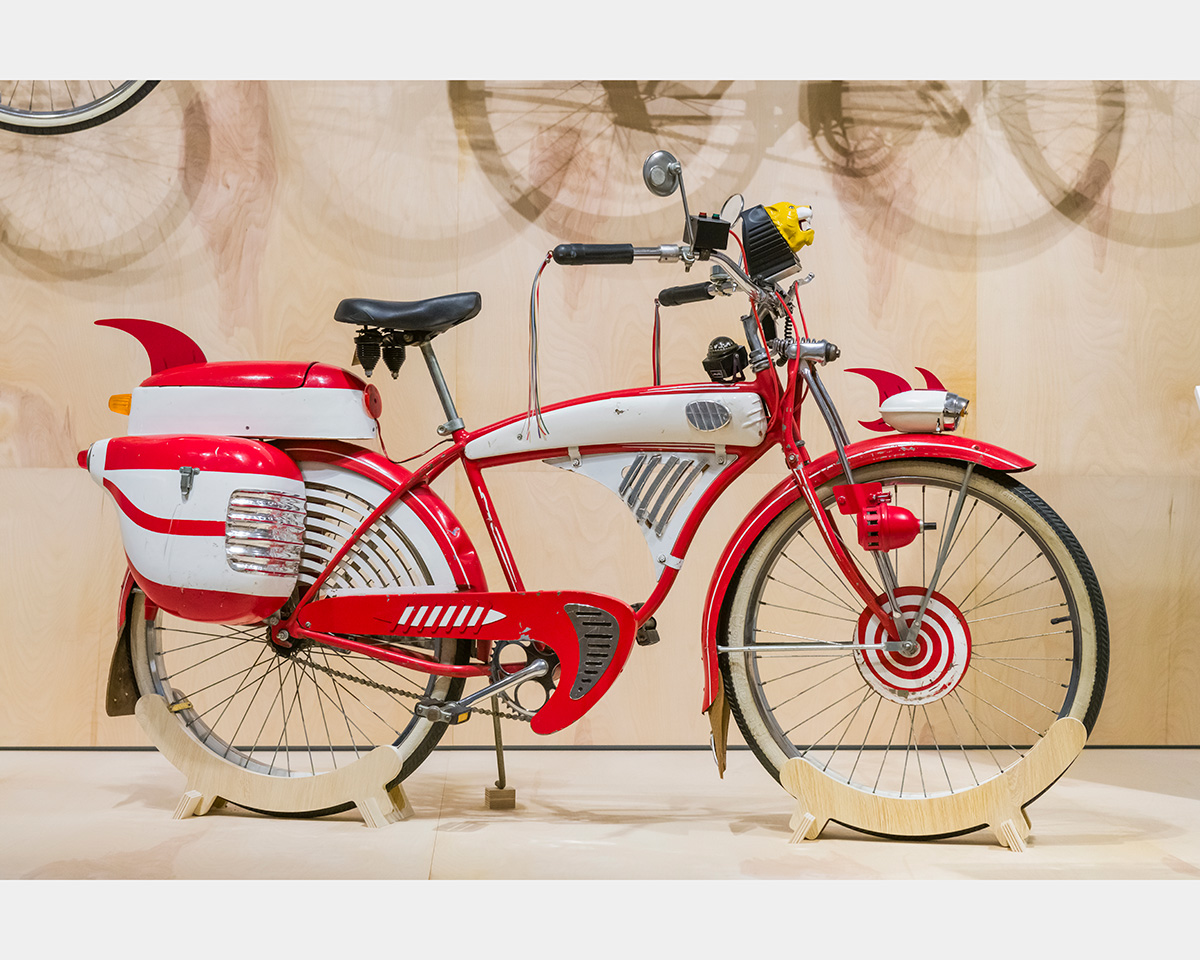 A red bicycle sits on a white floor. Shadows of bicycles are projected onto a tan-colored wall in the background.