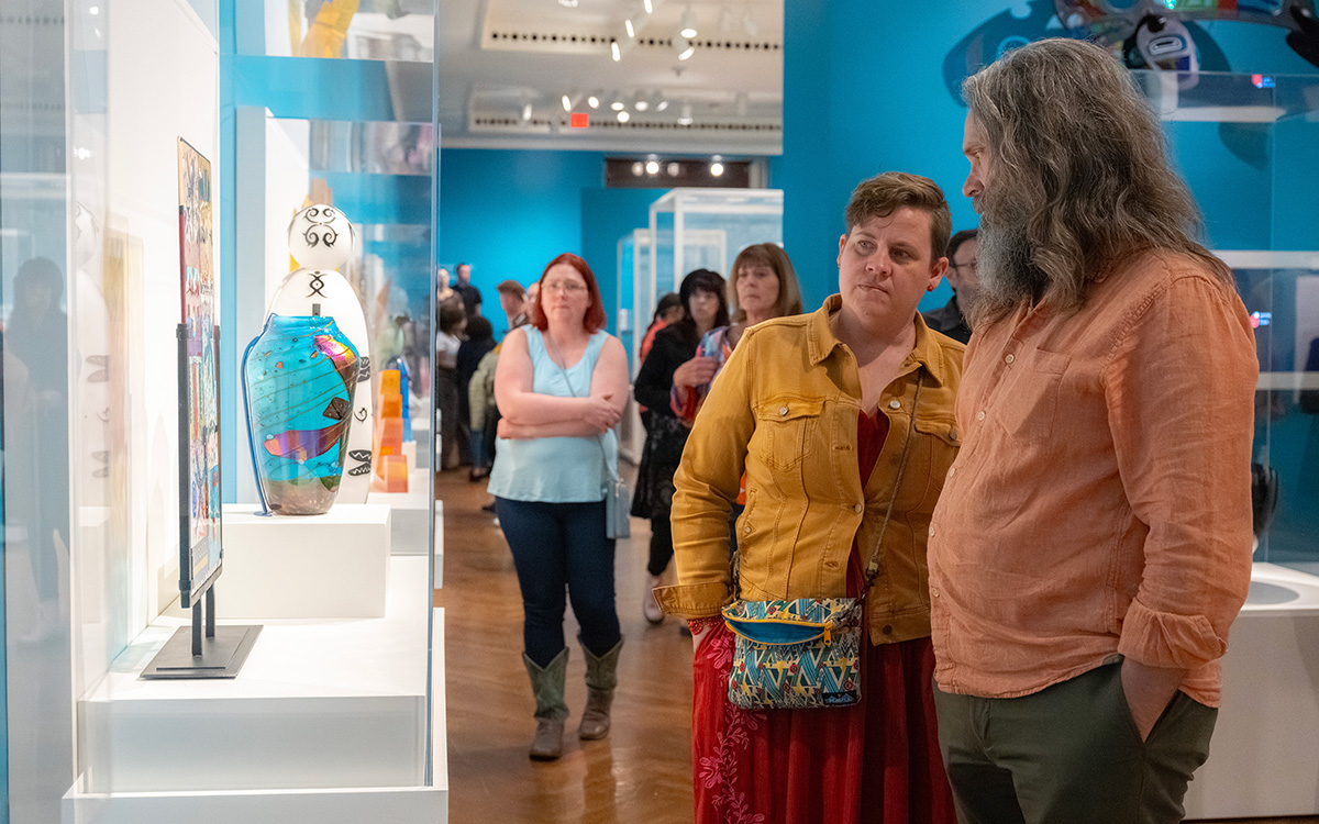 Two white visitors look at a bright blue glass vase in a gallery