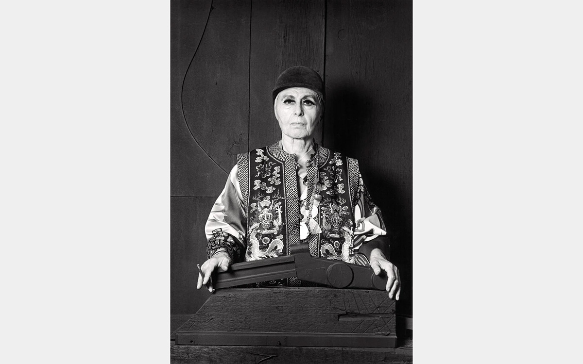 Black and white image of a woman dress in a cap and an elaborately patterned vest, standing behind an abstract sculptural arwork.