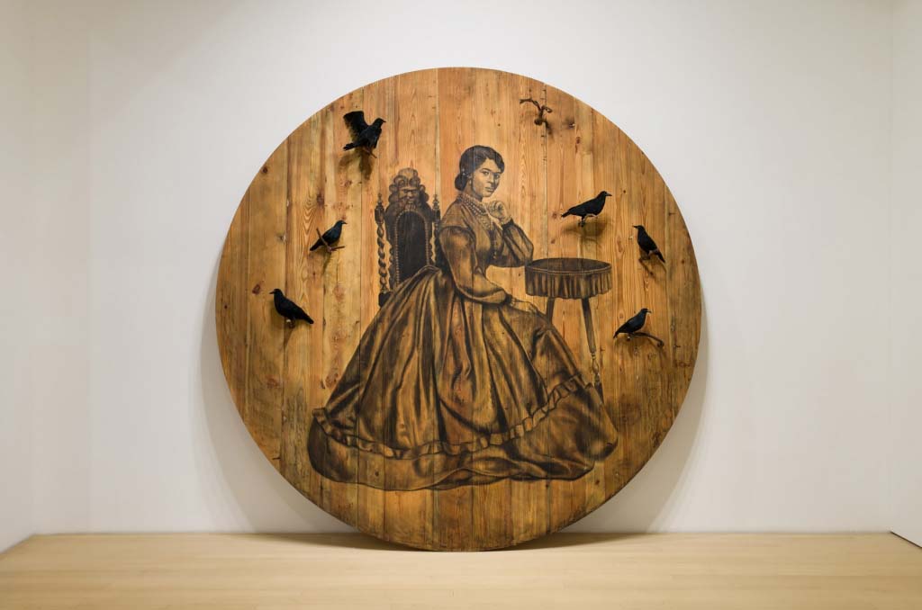 Large circular painting on wood plank, of a black woman dressed in a mid-19th century dress sitting at a small table. Several 3D crows are attached to the surface.