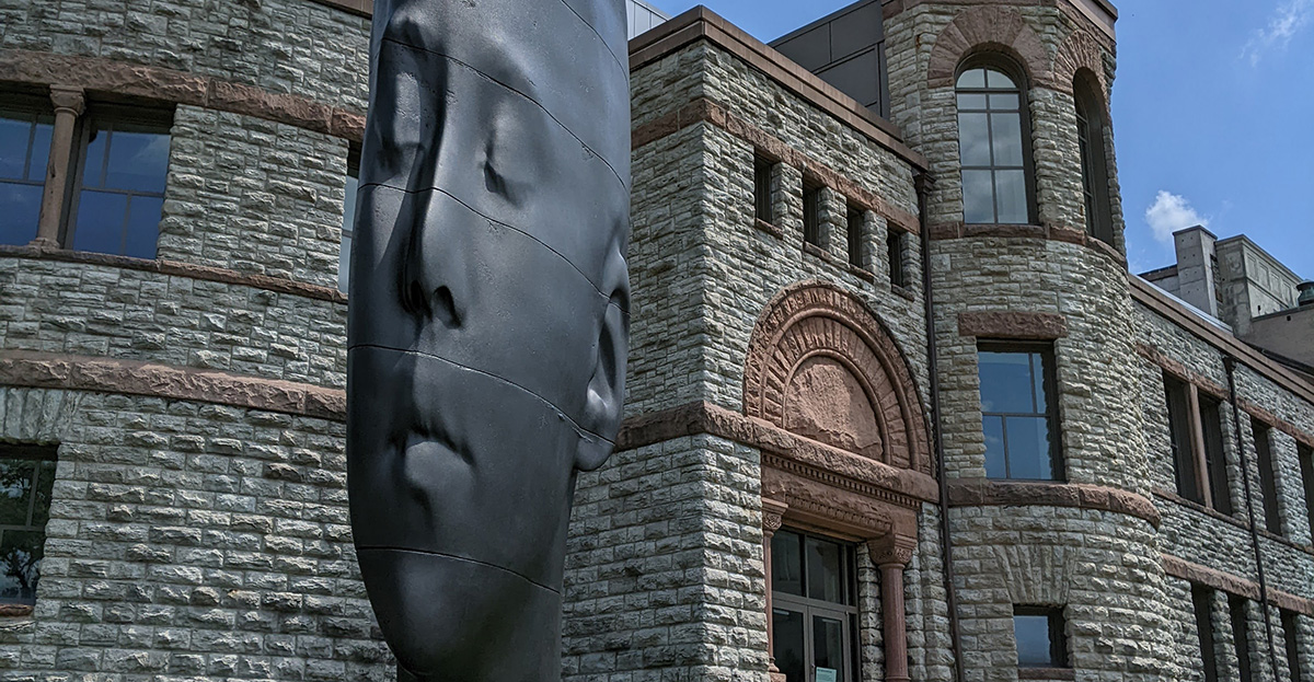 A monumental, thin black sculpture of a face in front of a stone building