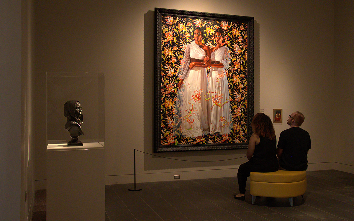 Two people sit on a yellow stool inside a gallery looking a painting depicting two women in white dresses.