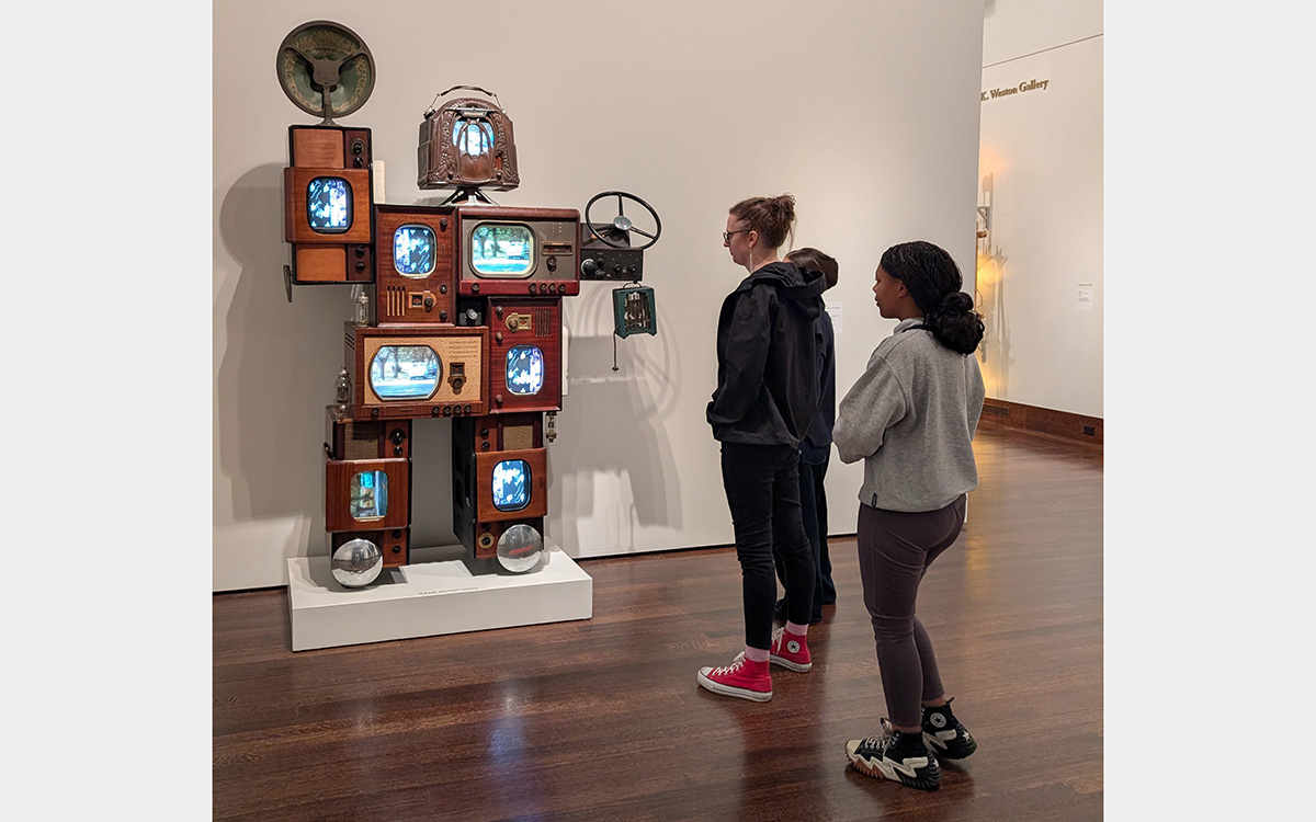 Three people looking at a robot-like figure composed of vintage television and radio sets and tech equipment